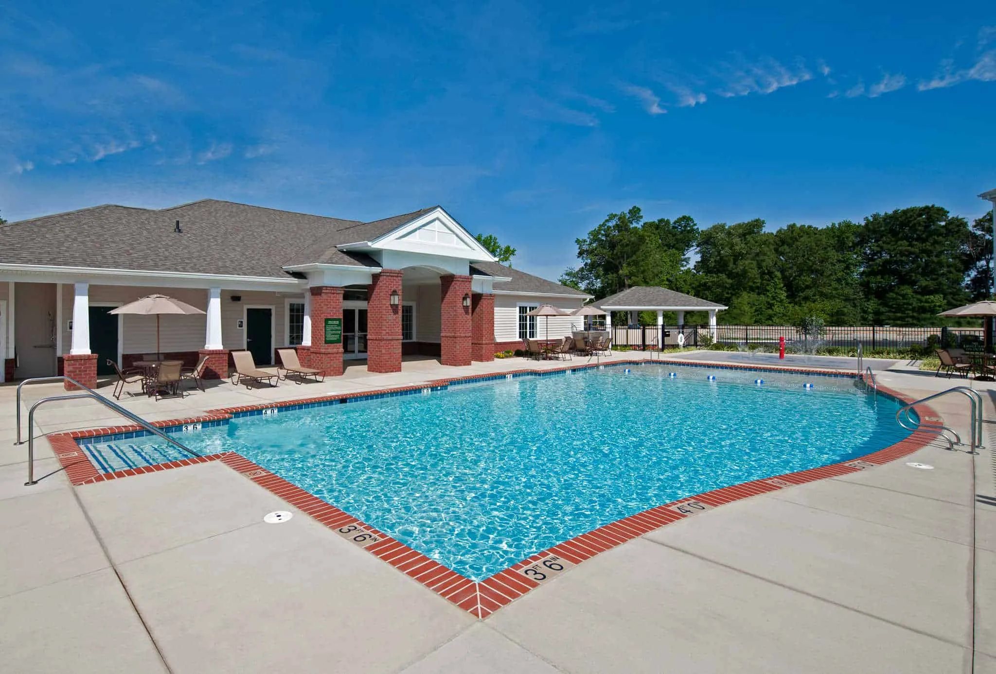 A large outdoor swimming pool with brick edging and a sundeck located next to a traditional clubhouse under a clear blue sky A large outdoor swimming pool with brick edging and a sundeck located next to a traditional clubhouse under a clear blue sky