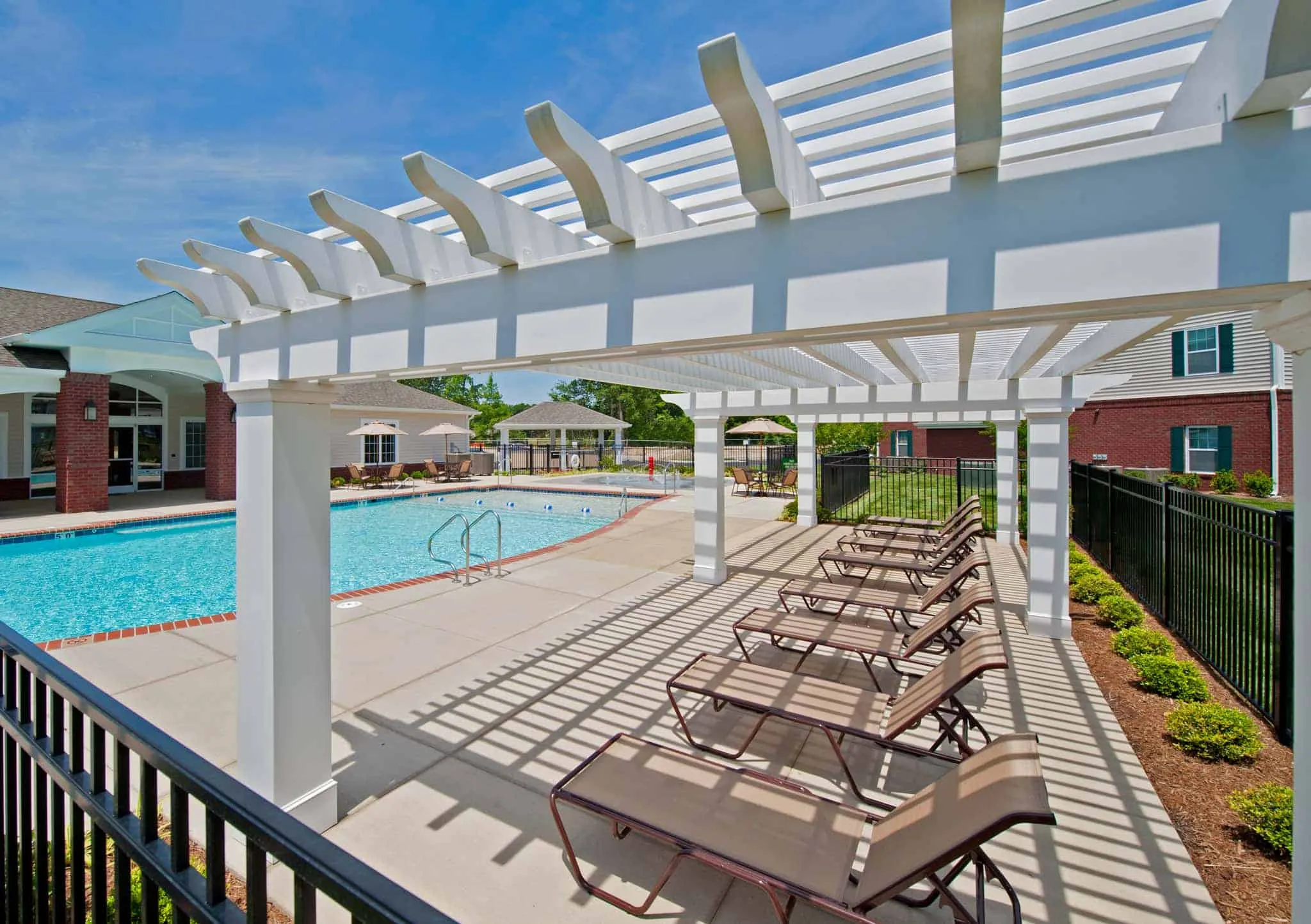 A view from underneath a white wooden pergola showing rows of lounge chairs on a concrete sundeck next to a swimming pool A view from underneath a white wooden pergola showing rows of lounge chairs on a concrete sundeck next to a swimming pool